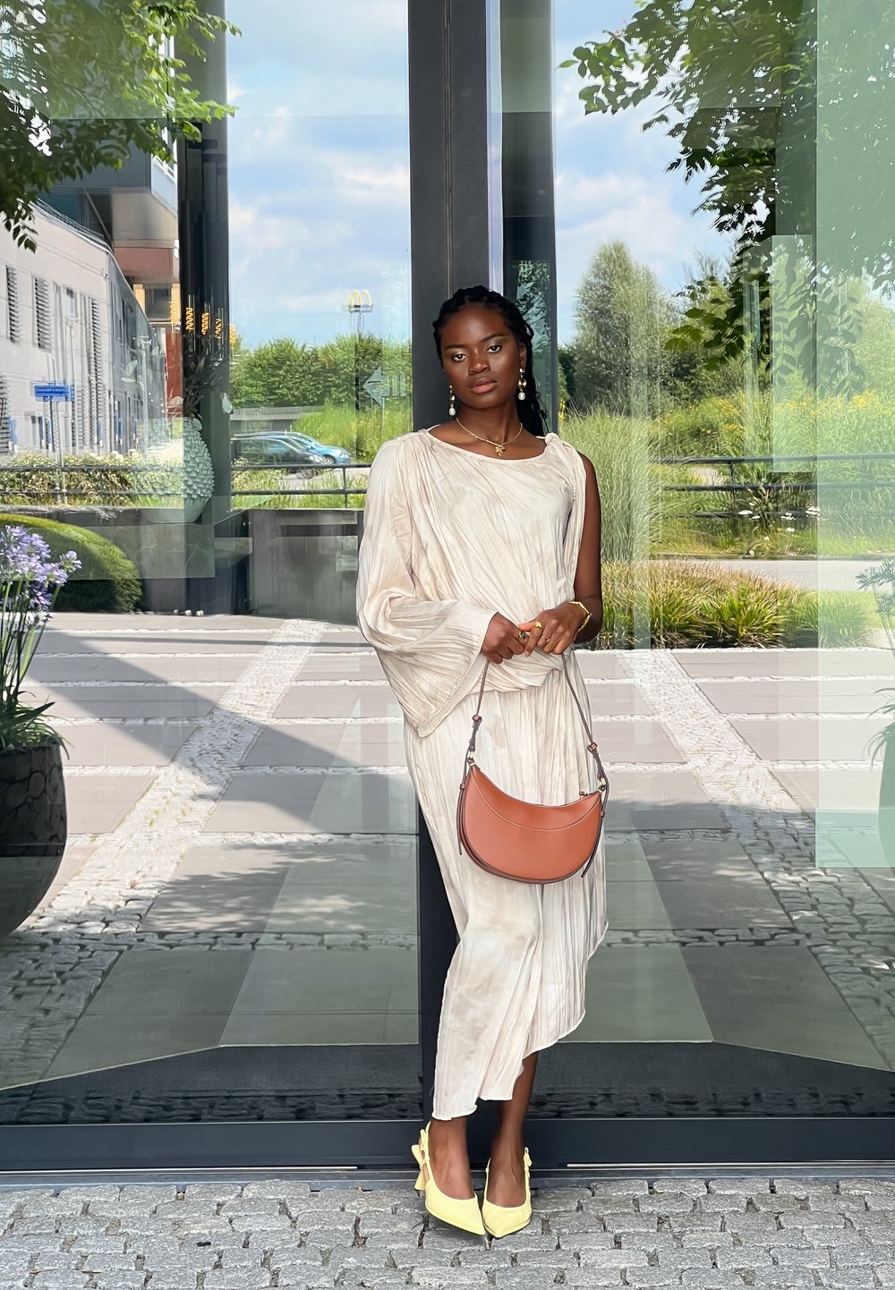 Robe créme fluide avec une manche asymétrique, assortie à un sac bandoulière marron et des talons jaunes. Texture lisse et arrière-plan de cadre naturel.