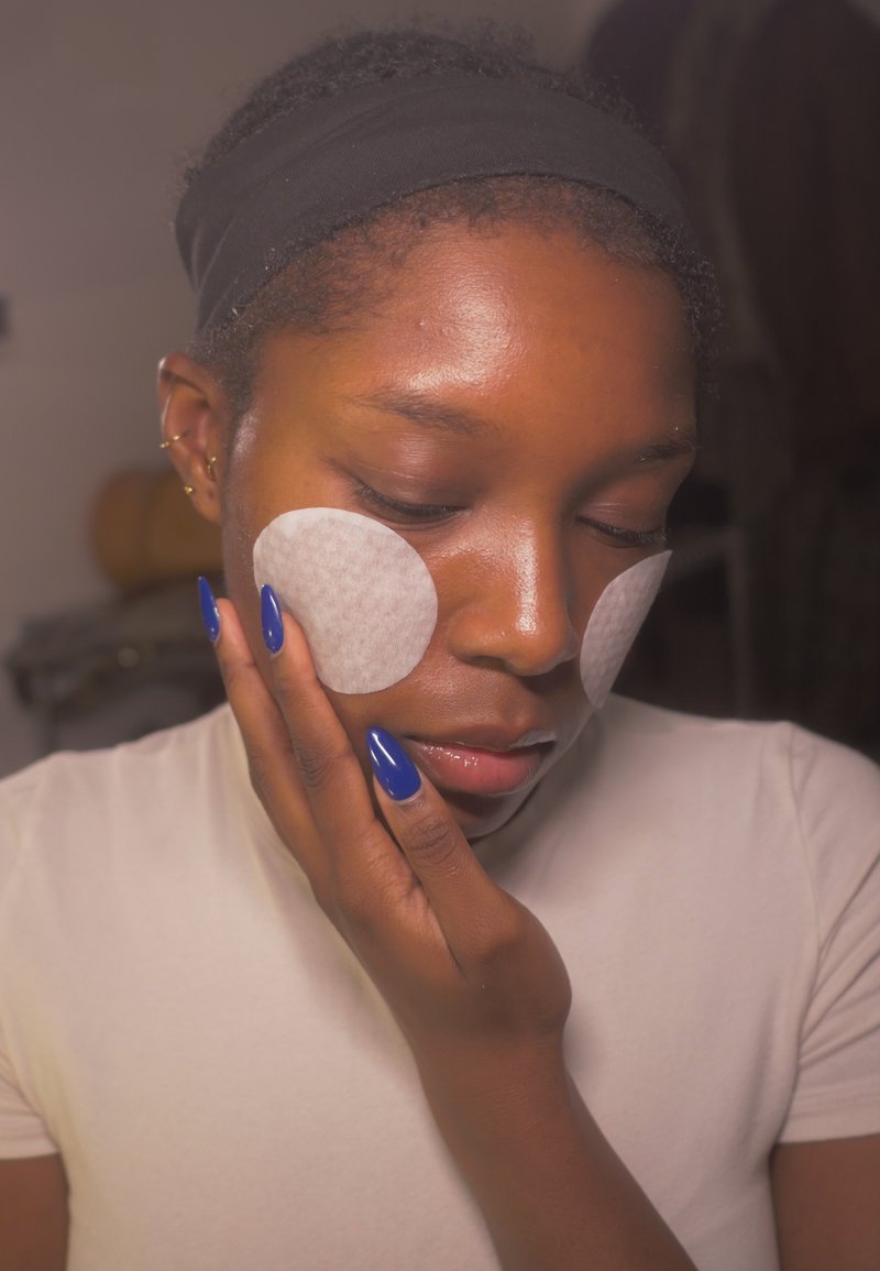 White round facial pads on cheeks, blue long nails on hand, light skin tone, wearing a fitted light shirt and black headband.