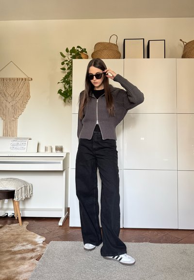 Mujer ajustándose unas gafas de sol negras, vestida con un suéter gris con cremallera, pantalones anchos negros y zapatillas blancas, en una habitación con armarios blancos y un piano.