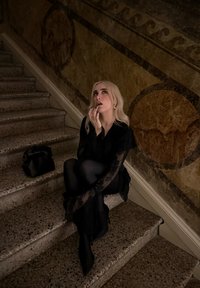 Femme blonde en robe noire en dentelle assise sur des marches en pierre, regardant pensivement vers le haut, à côté d'un sac à main noir et d'une décoration murale ornée.