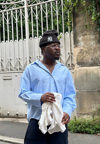 Ανοιχτό μπλε πουκάμισο με κουμπιά, μαύρη σκούφη με λογότυπο, κρατάει λευκό ύφασμα, στέκεται κοντά σε διακοσμητική πύλη, φοράει γυαλιά και κοσμήματα.