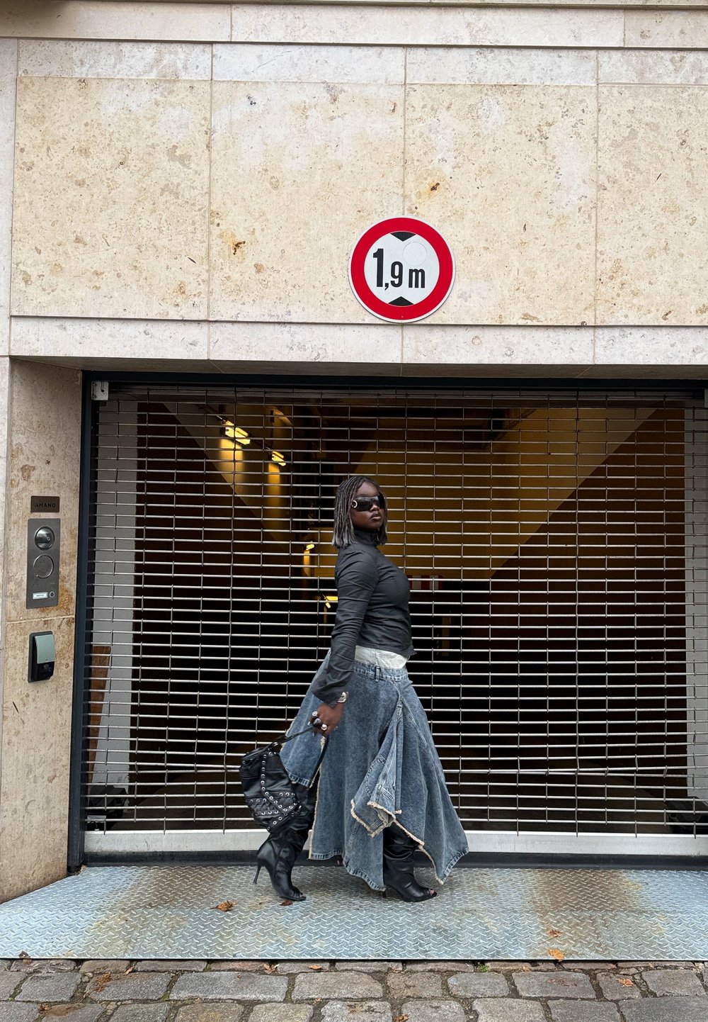 Čierna košeľa s dlhým rukávom, rifľová sukňa s ošúchanými okrajmi, čierne topánky po kolená a čierna kabelka. Pred kovovou mrežou a výškovou tabuľou.