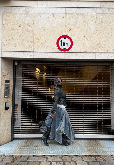 Camisa negra de manga larga, falda vaquera con bordes deshilachados, botas negras hasta la rodilla y un bolso negro. Frente a una rejilla metálica y un cartel de altura.