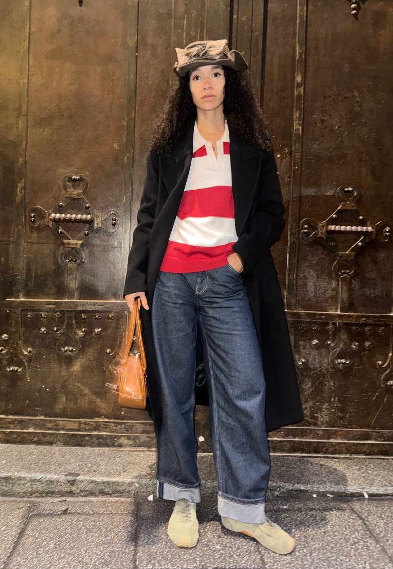 Femme aux cheveux bouclés portant un chapeau marron, un manteau noir, une chemise rayée rouge et blanche, un jean large, des chaussures beige, tenant un sac à main marron.