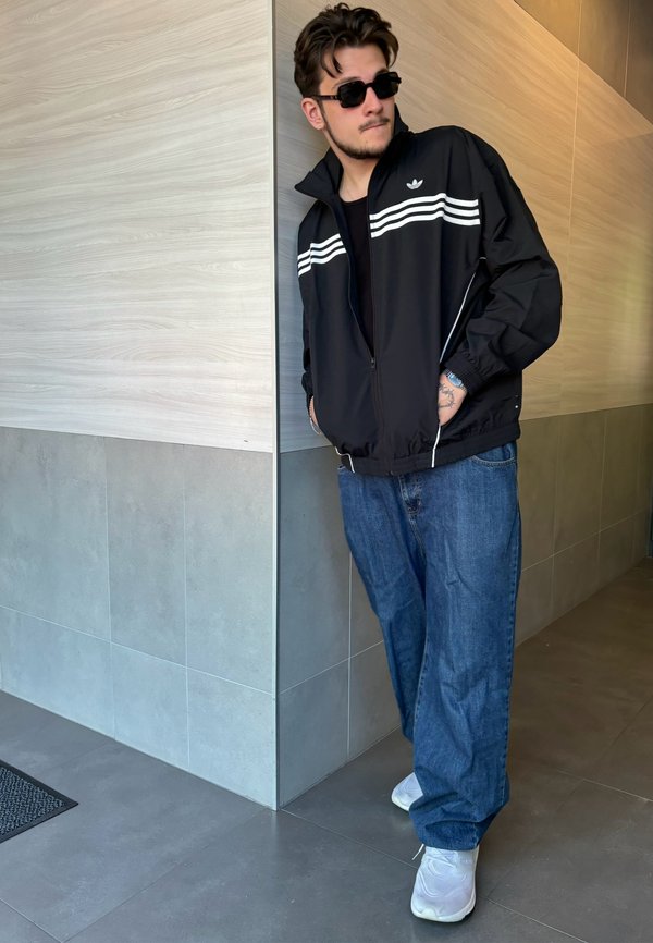 Casaco atlético preto com riscas brancas, jeans de ganga azuis, e ténis brancos. O casaco possui fecho zipper e bolsos laterais.