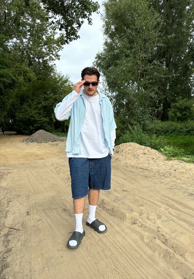 Camisa de botón azul claro sobre una camiseta blanca, pantalones cortos de mezclilla, zapatos negros sin cordones y calcetines blancos. Sendero de arena y vegetación de fondo.