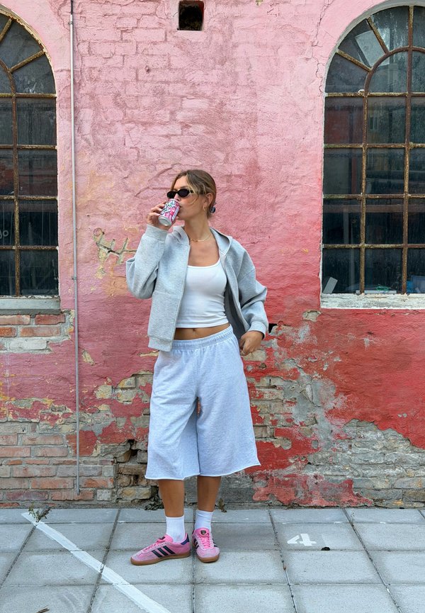Jeune femme en shorts de jogging gris et veste boit dans une canette, debout contre un mur de briques roses usé avec deux fenêtres en arc.