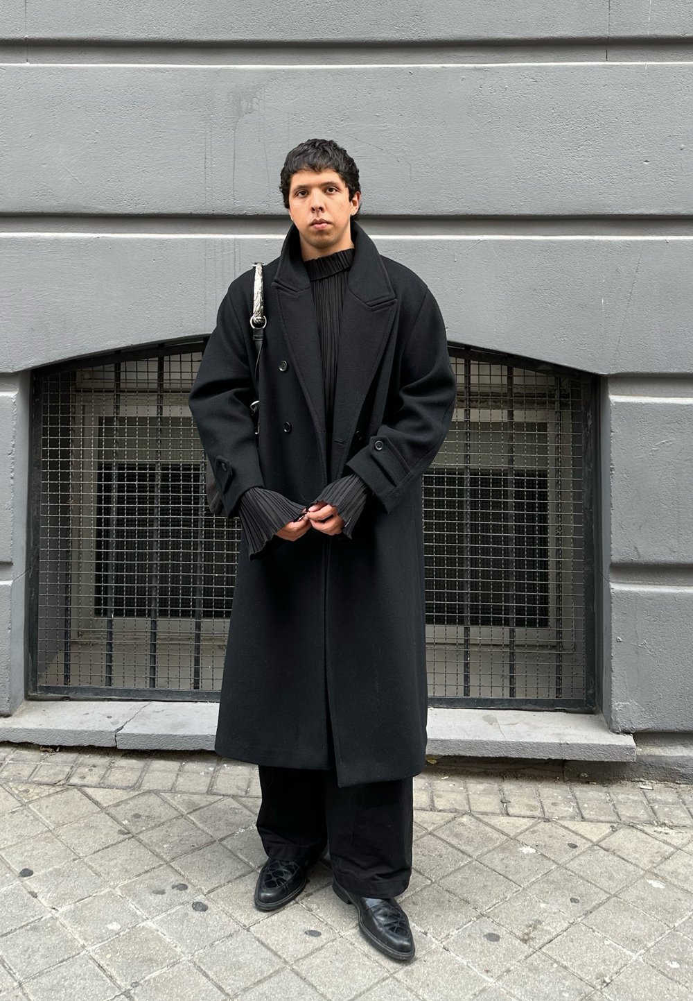 Μαύρο παλτό διπλής όψης με φαρδιές πέτες και πτυχώσεις στους μανικέτες, φορεμένο πάνω από μια ριχτή μαύρη μπλούζα και φαρδιές μαύρες παντελόνες, με μαύρα παπούτσια.