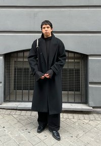 Μαύρο παλτό διπλής όψης με φαρδιές πέτες και πτυχώσεις στους μανικέτες, φορεμένο πάνω από μια ριχτή μαύρη μπλούζα και φαρδιές μαύρες παντελόνες, με μαύρα παπούτσια.