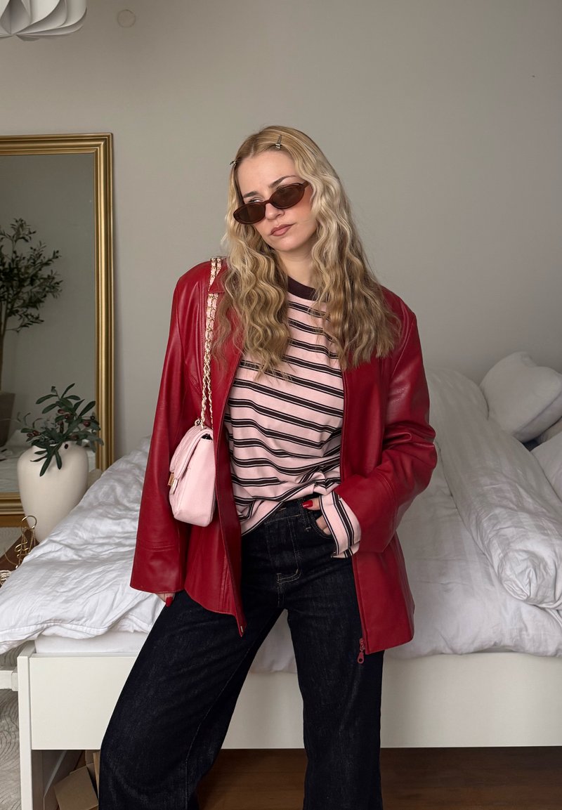 Femme aux longs cheveux blonds ondulés portant des lunettes de soleil, une veste en cuir rouge, une chemise rayée rose, un jean noir et un sac rose, debout dans une chambre.