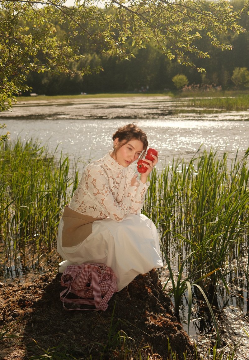 Joven mujer con blusa de encaje blanca y falda agachada cerca de un lago, sosteniendo dos pimientos rojos cerca de su cara, con la luz del sol reflejándose en el agua.