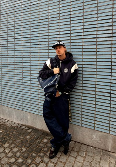 Joven con sudadera negra y gorra apoyado contra una pared de azulejos azules, sosteniendo un bolso de lona azul marino en una acera de adoquines.