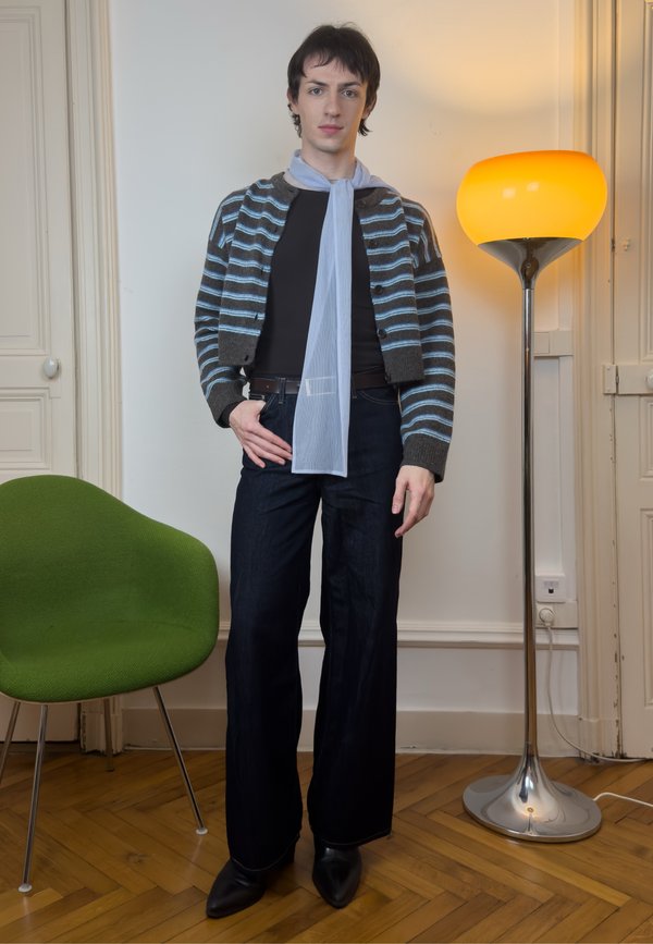 Persoană tânără cu cardigan cu dungi, top negru, pantaloni cu picioare largi și eșarfă albastru deschis stând în interior lângă un scaun verde și o lampă de podea.