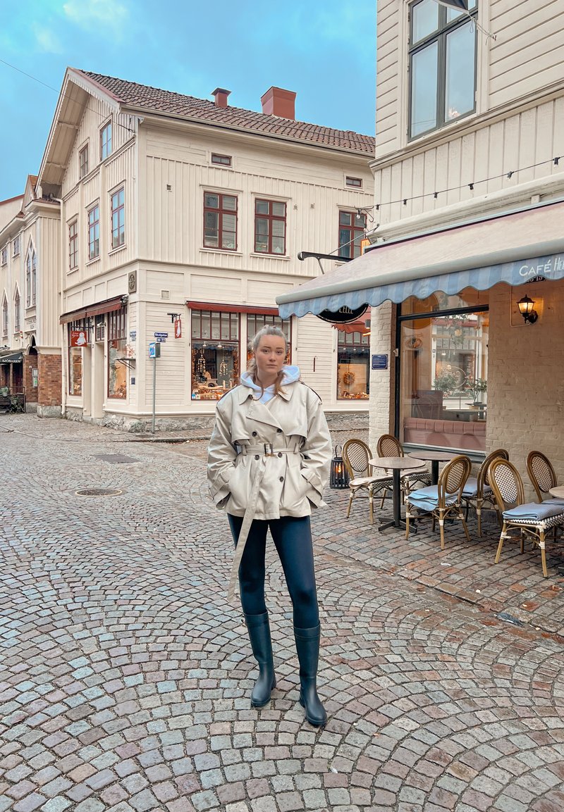 Kvinde iført beige trenchcoat og sorte støvler, stående på brostensbelagt gade nær udendørs caféborde og træbygninger.