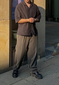 Μαύρο κοντομάνικο πουκάμισο, γκρι χαλαρές παντελόνες και χοντρά μαύρα παπούτσια. Υφασμάτινη υφή με διακριτό μοτίβο. Στηρίζει το σώμα του σε έναν τοίχο από πέτρα.