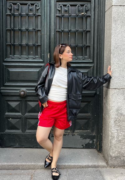 Chaqueta de piel sintética negra sobre una blusa blanca, combinada con shorts deportivos rojos con franjas blancas. Ballet flats negros de punta con tachuelas.