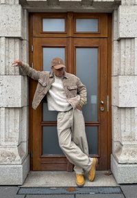 Homme en veste beige, casquette et pantalon appuyé contre un mur en pierre près d'une porte en bois, une main dans la poche et les jambes croisées.