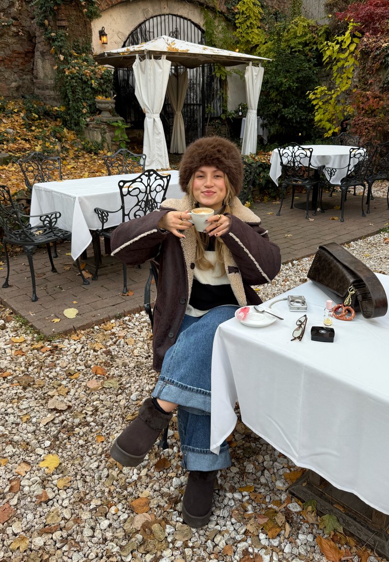 Femme portant un chapeau et un manteau en fourrure assise à une table de café en plein air, tenant une tasse de café, des feuilles d'automne éparpillées au sol.