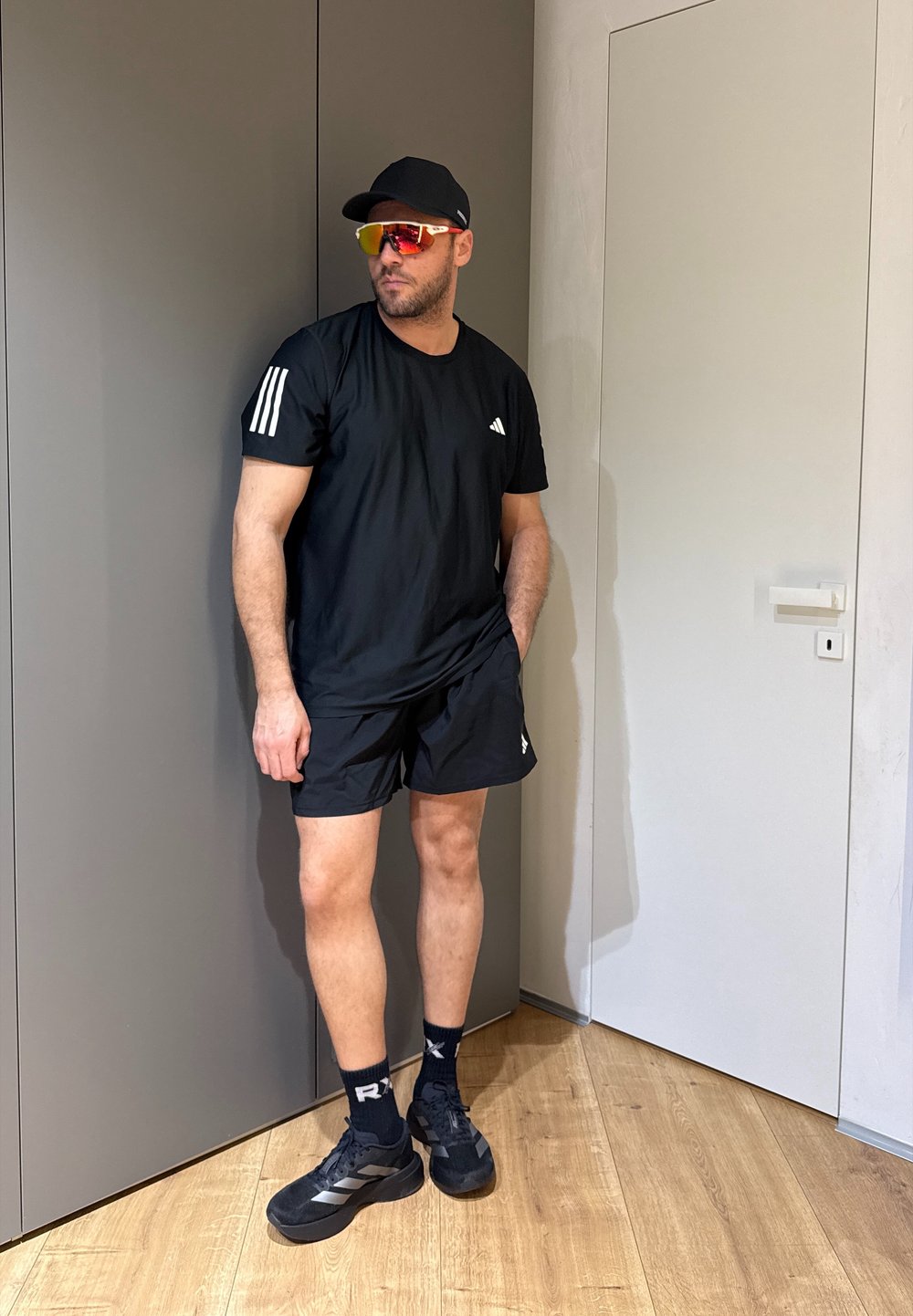 Homme portant une tenue de sport noire, une casquette noire et des lunettes de sport réfléchissantes, debout à l'intérieur près d'un mur gris et d'une porte blanche.