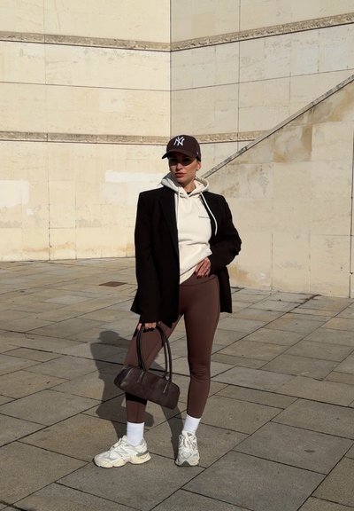 Mujer con leggings marrones, sudadera blanca con capucha, blazer negro, zapatillas deportivas y una gorra de béisbol oscura, de pie al aire libre sosteniendo un bolso marrón.