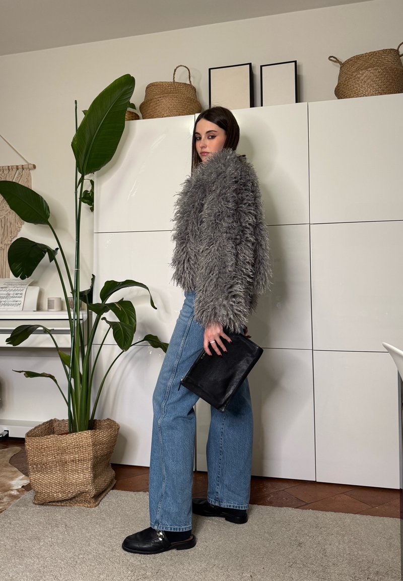 Frau in blauer Jeans und grauer flauschiger Jacke hält eine schwarze Clutch und steht neben einer großen grünen Topfpflanze in einem minimalistischen Raum.