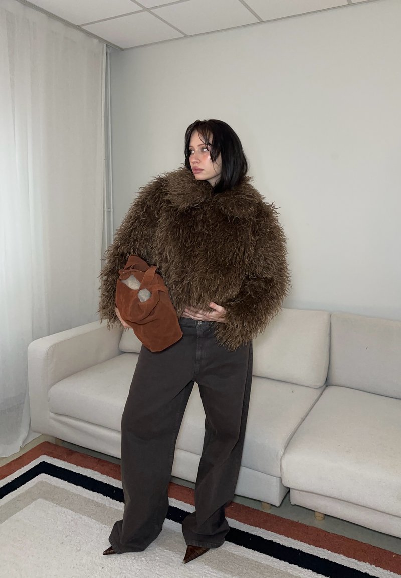 Femme portant une veste marron duveteuse, un pantalon large sombre, des chaussures à bouts pointus, tenant un sac en daim marron, debout à côté d'un canapé blanc dans une pièce minimaliste.