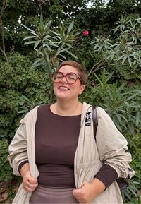 Persona con capelli corti e grandi occhiali rossi che sorride, indossando una giacca beige e una maglia scura, in piedi di fronte a piante verdi folte.