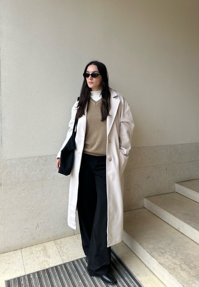 Suéter de lana beige superpuesto sobre un cuello de tortuga blanco, combinado con pantalones anchos negros y un abrigo largo beige. Bolsa de tote negra y gafas de sol.