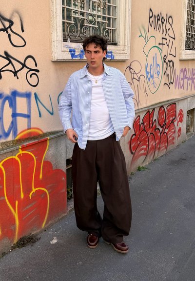 Joven con camisa a rayas, camiseta blanca y pantalones anchos marrones de pie en una calle urbana cubierta de grafitis junto a una pared beige con ventanas con rejas.