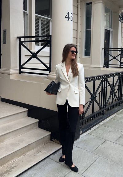 Blazer blanco sobre pantalones negros, sosteniendo un bolso negro. Ella está de pie en unos escalones de piedra, con un edificio de ladrillo y una barandilla decorativa al fondo.