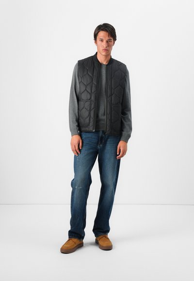 Conjunto de hombre que incluye un chaleco acolchado negro sobre un suéter gris, combinado con jeans azules y zapatos marrones claros. Diseño simple y colores neutros.