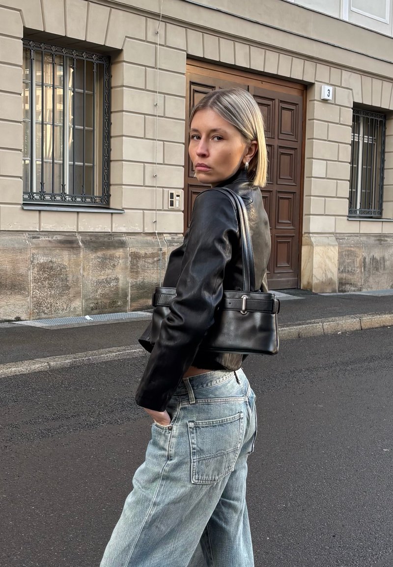 Young woman in black leather jacket and light blue jeans stands on street, carrying black handbag over shoulder, looking back toward camera.