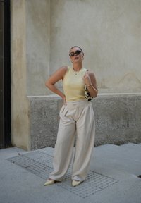 Hellgelbes, geripptes, ärmelloses Oberteil, cremefarbene, gefaltete, weite Hose, spitze beigefarbene Schuhe, schwarze Sonnenbrille und eine gemusterte Handtasche.
