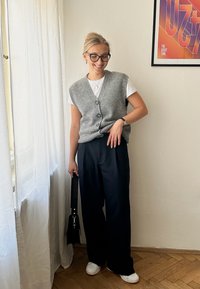 Grå ermestløse kardigan over en hvit t-skjorte, paret med mørke vide bukser og hvite slip-on sko, står nær gjennomsiktige gardiner.