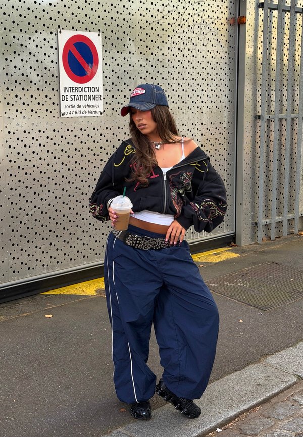 Joven mujer con pantalones anchos, sudadera corta con capucha y gorra, sosteniendo café con hielo, de pie en la calle cerca de una puerta de metal y una señal de prohibido estacionar en francés.