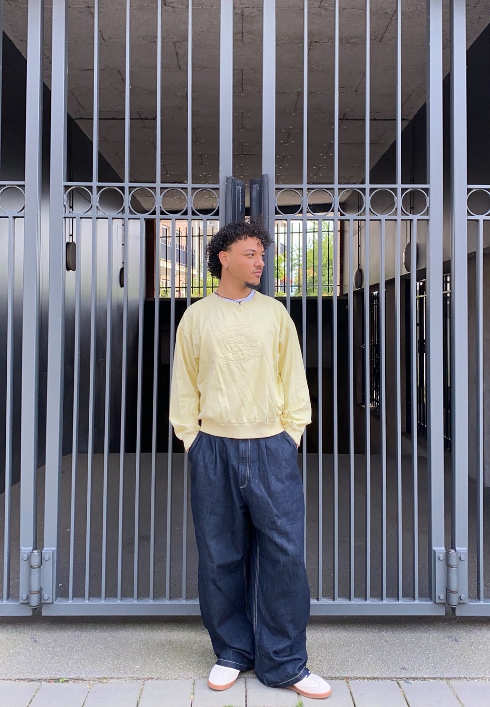 Sweater amarelo claro com bordado frontal, calças de ganga escuras e largas, e sapatilhas brancas contra um portão vertical cinza.