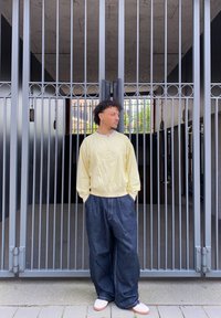Sweater amarelo claro com bordado frontal, calças de ganga escuras e largas, e sapatilhas brancas contra um portão vertical cinza.