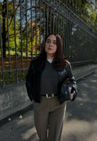 Zwarte leren jas over een grijze trui, gecombineerd met hooggesneden beige broeken. Een kleine zwarte handtas maakt de outfit compleet.