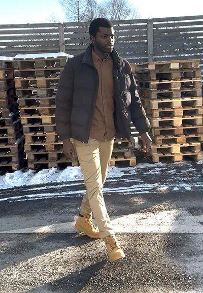 Hombre con chaqueta marrón, pantalones color caqui y botas caminando sobre un pavimento nevado junto a palets de madera apilados y una cerca de madera.