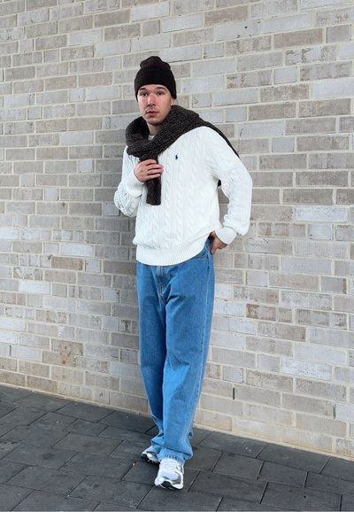 Jersey blanco de punto tipo cable, vaqueros azules de corte holgado, bufanda marrón y gorro a juego. Zapatillas blancas con detalles grises, posando frente a una pared de ladrillos.