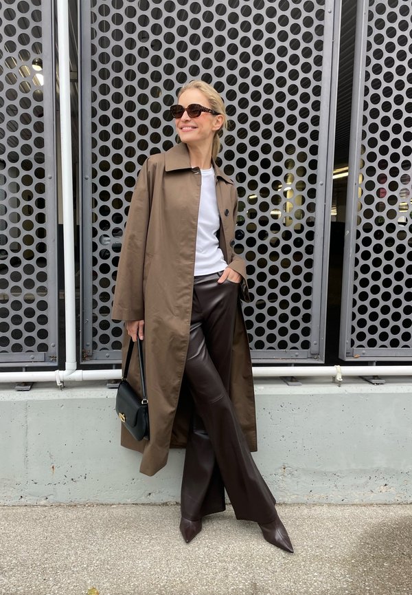 Brown trench coat over a white shirt, paired with brown faux leather pants and pointed brown heels. Holds a black handbag.
