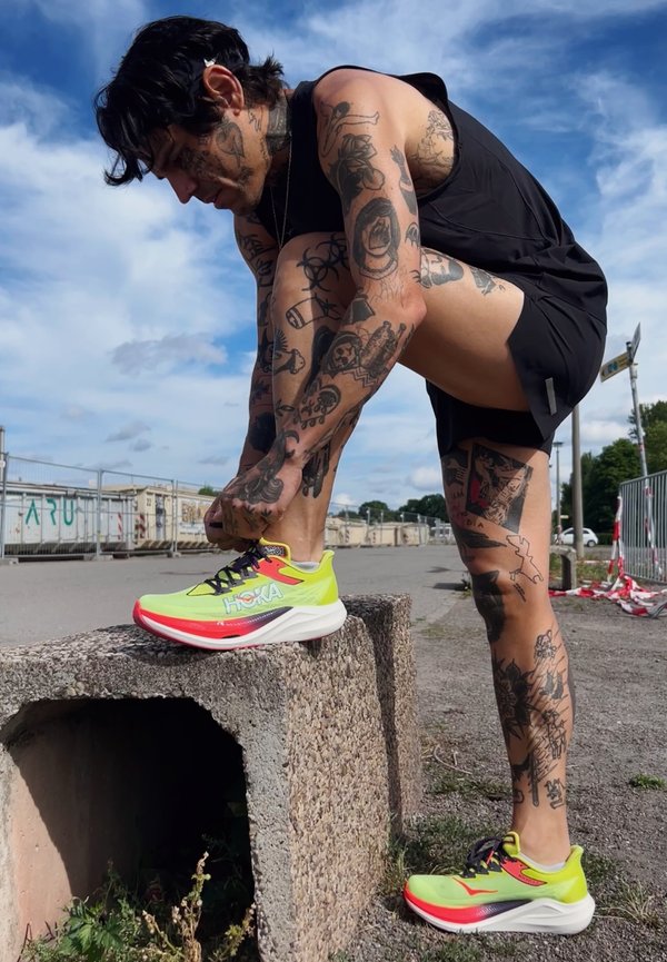 Persoană tatuată în îmbrăcăminte sport neagră îngenunchează în aer liber, legându-și adidașii Hoka galben neon pe un bloc de beton, cu cer albastru pe fundal.