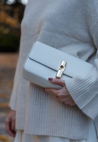 Bijela kožna torbica s glatkom teksturom, pravokutnog oblika, s kopčom u zlatnom tonu. Torbica ima jednostavan, čist dizajn.