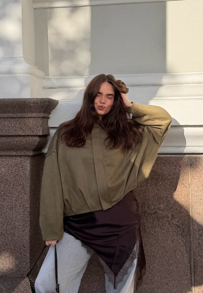 Joven mujer con cabello largo y castaño con chaqueta oliva oversize y falda negra sobre pantalones blancos posa frente a una pared de piedra y blanca, sonriendo con los ojos cerrados.