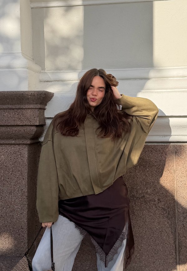 Jonge vrouw met lang bruin haar in een oversized olijfgroene jas en zwarte rok over witte broek poseert tegen een stenen en witte muur, glimlachend met gesloten ogen.