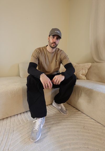 Hombre con gorra marrón claro, camiseta marrón claro sobre mangas largas negras, pantalones negros y zapatillas grises sentado en un sofá seccional beige en una habitación de tonos neutros.