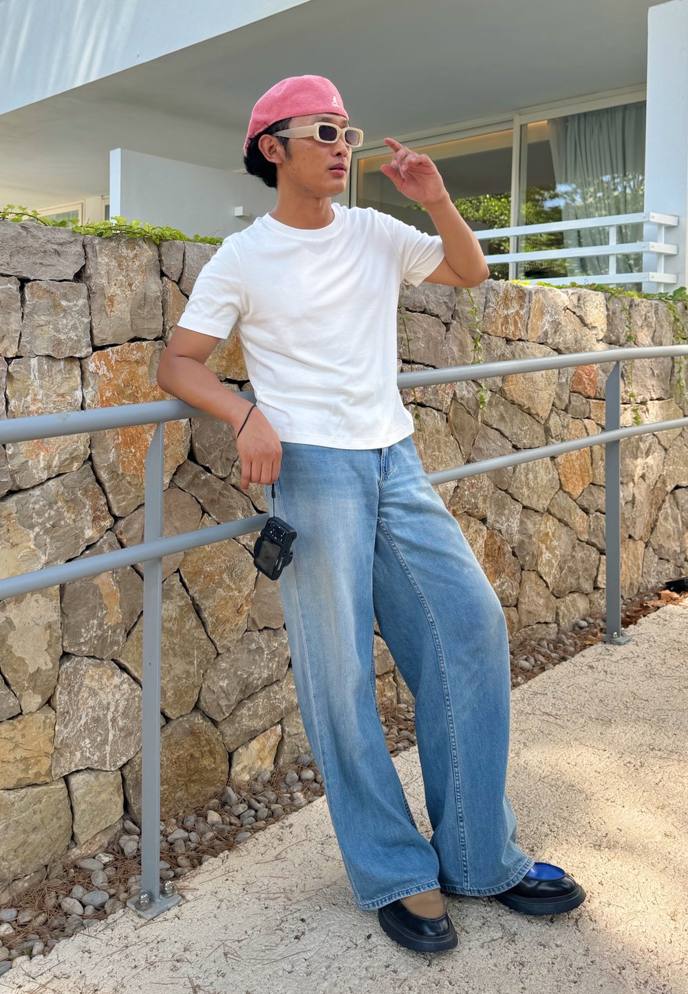Wijdvallende, lichtblauwe denim jeans, wit shirt met korte mouwen, roze pet en oversized zonnebril. Een compacte camera hangt aan de tailleband.