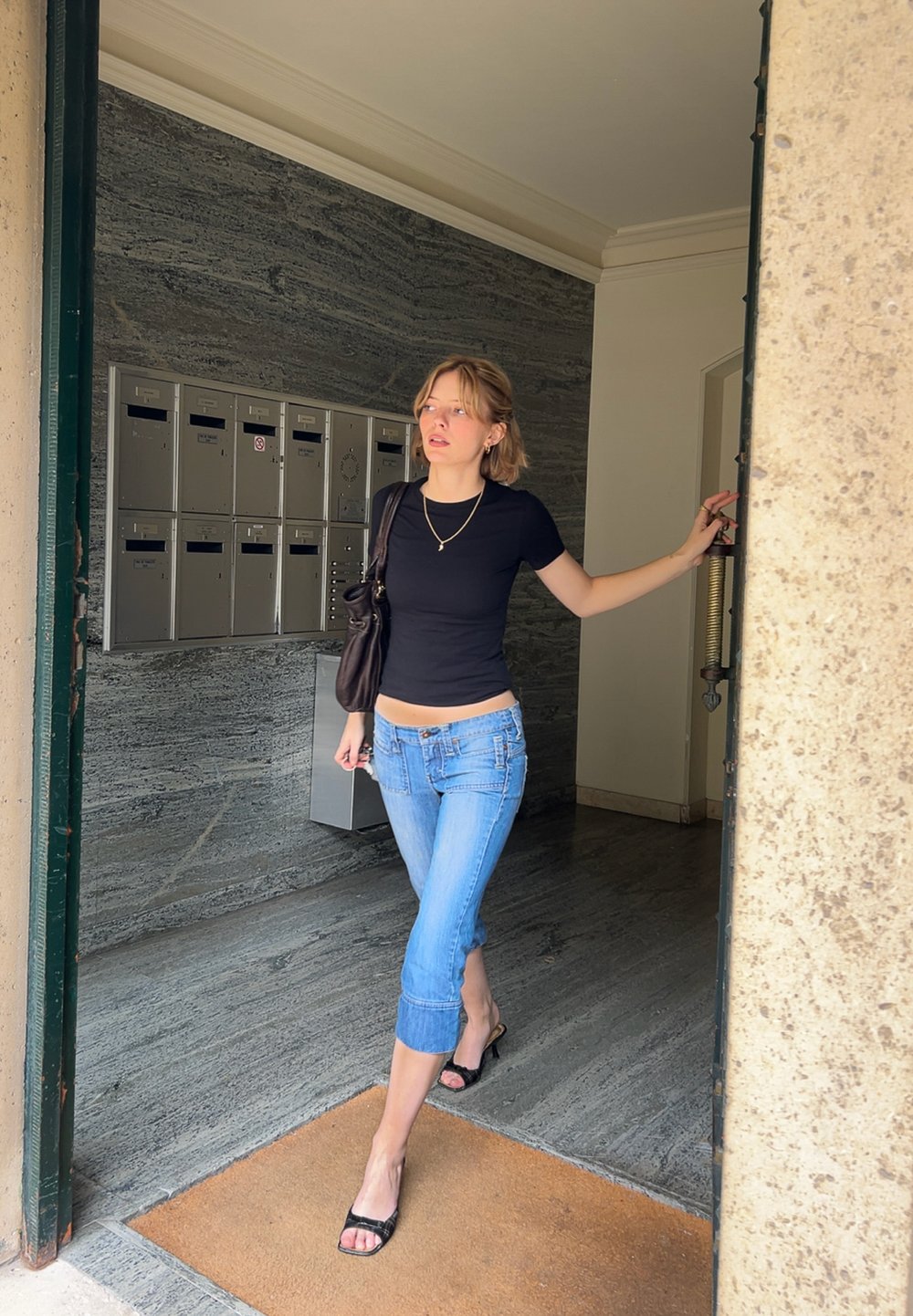 Schwarzes Kurzarmoberteil, hellblaue, cropped Jeans, schwarze High-Heels, goldene Halskette. Innenbereich mit Briefkästen und Steinmauer.