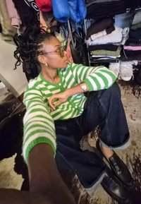 Žaliai ir baltai dryžuotas megztinis kardiganas, džinsai su užlenktais kulnais, juodi loafer batai, o fone matyti netvarkingas drabužių stovas.