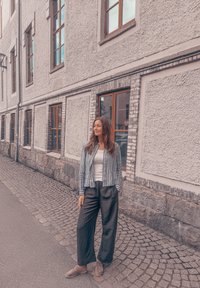 Camisa de botão riscada, top branco de alças, calças largas pretas e sapatos castanhos. Rua de calçada e paredes de pedra texturizada com janelas.